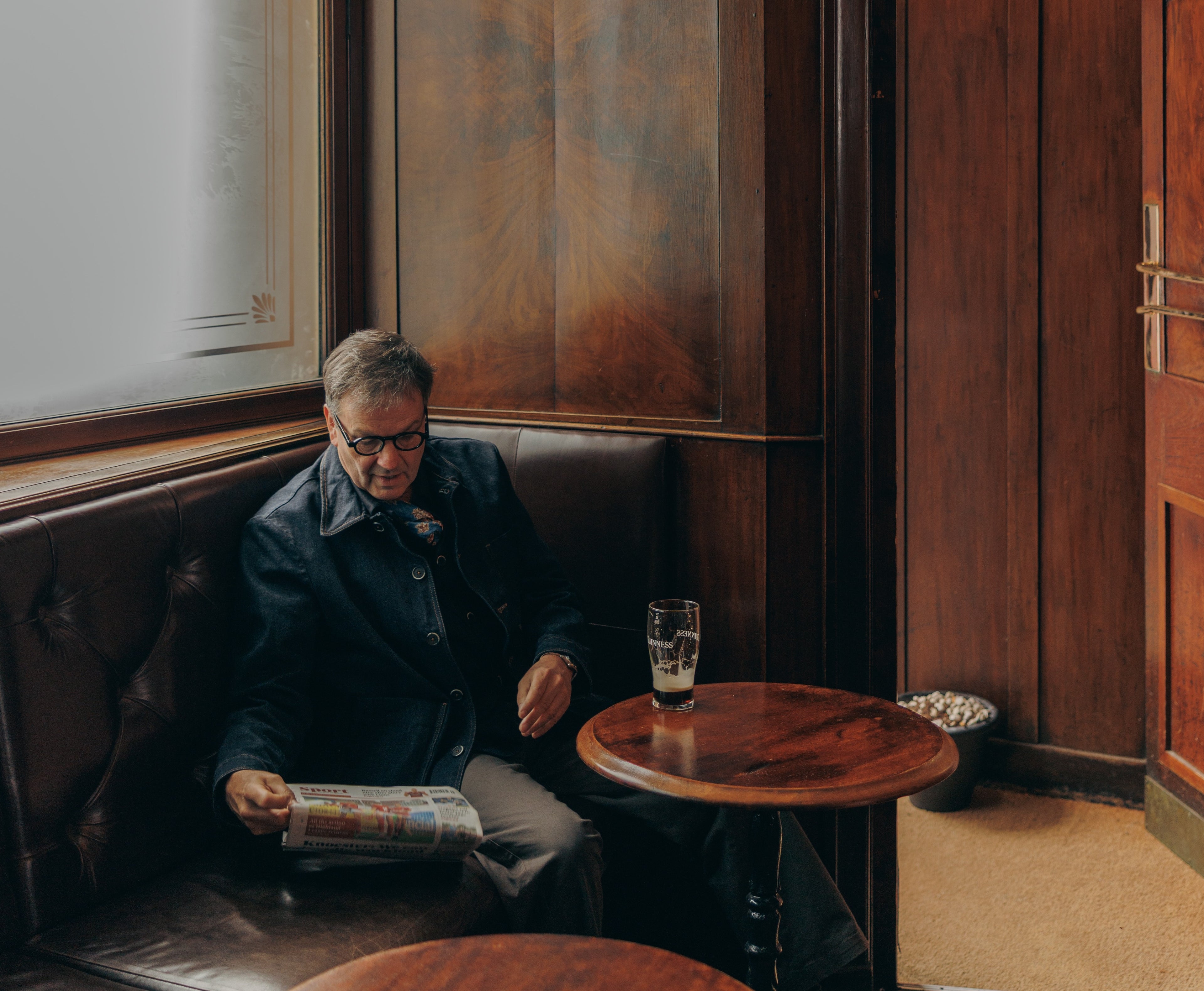 Man sitting in a dimly lit bar with a drink and newspaper wearing Barena of Venezia.