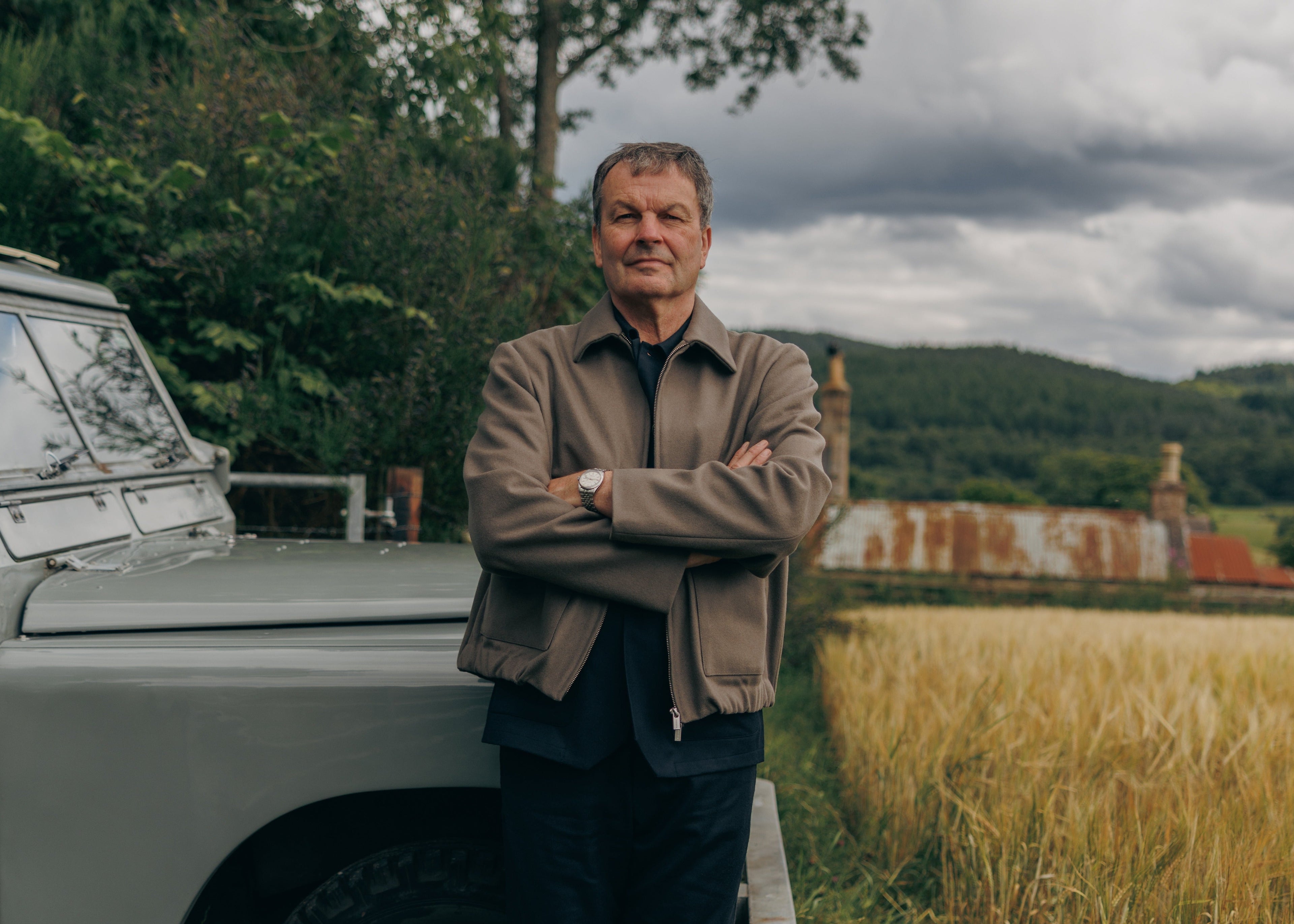 Man standing next to a vintage Land Rover in a rural setting with fields and trees.