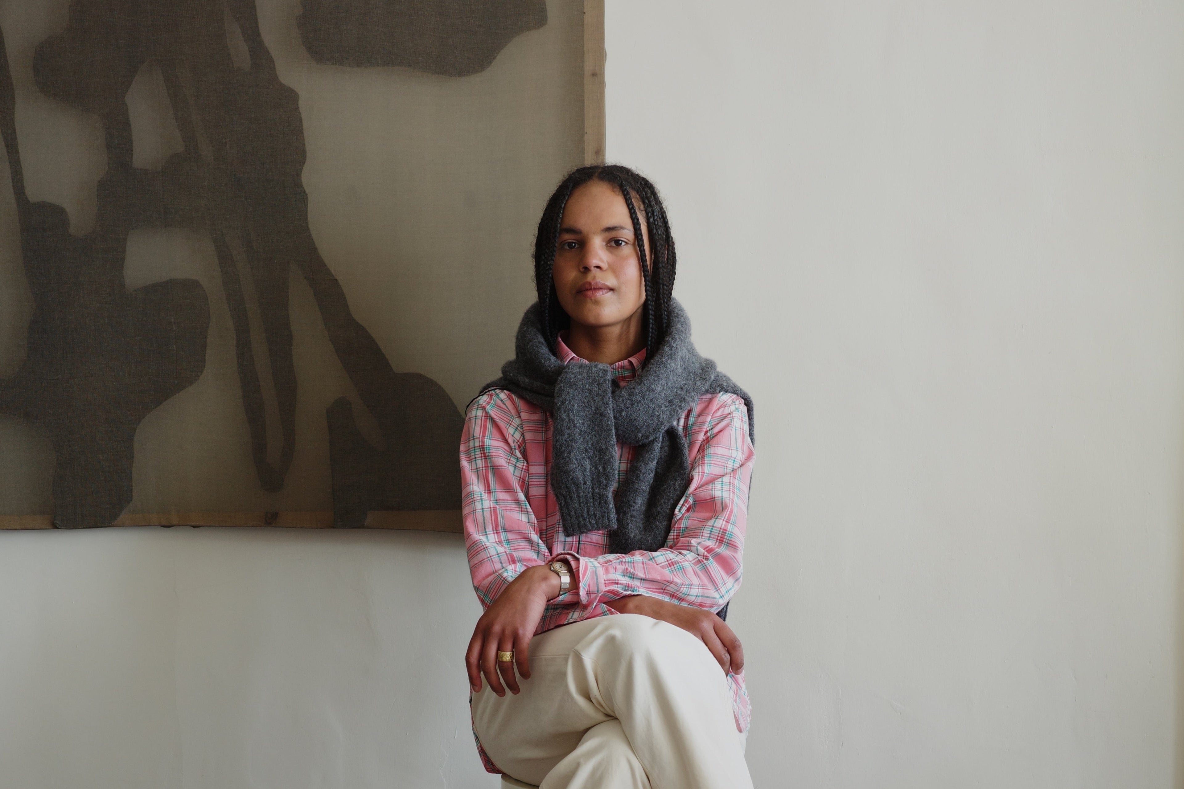 Woman sitting on a chair wearing a plaid shirt, gray scarf, white pants, and black shoes in a room with a textured wall.